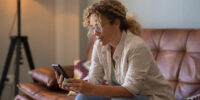 Serious young woman text messaging using mobile phone while sitting on sofa at home. Woman with curly hair and eyeglasses reading or texting using mobile phone in the living room